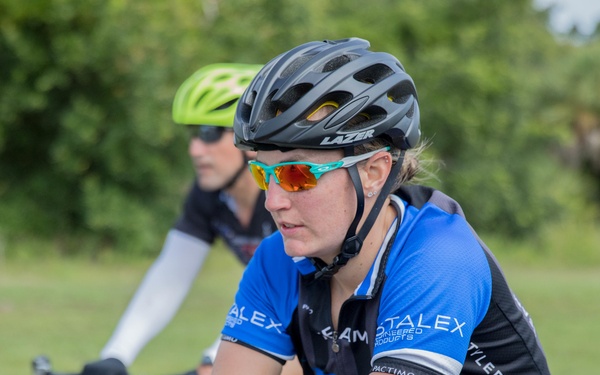 Two cyclists are riding on a paved road surrounded by green vegetation. The foreground cyclist, a woman, is wearing a black Lazer helmet, red-mirrored cycling sunglasses, and a blue and black team jersey reading 'STALEX' and 'Tyler Hamilton Training.' A second cyclist is visible in the background wearing a yellow-green helmet.