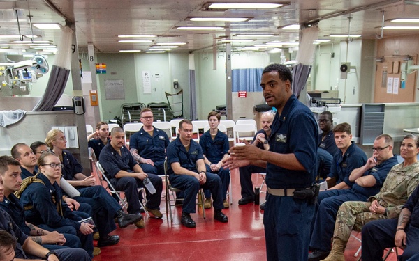 A military briefing or training session is taking place in what appears to be a shipboard medical bay or sick bay facility. A standing individual in navy blue coveralls addresses a large group of seated personnel, most wearing matching navy blue coveralls with some in camouflage uniforms. The setting features medical equipment, surgical lights, and a distinctive red floor consistent with a naval vessel's medical department.
