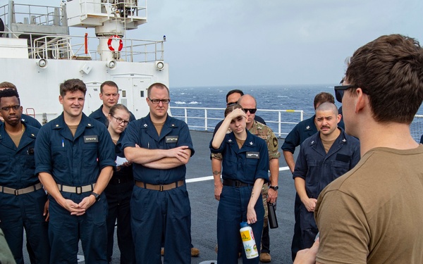 A group of approximately 15-20 military personnel in dark navy blue uniforms are gathered on the deck of a naval vessel at sea, listening to a briefing from an individual in an olive/tan t-shirt and green trousers. The setting appears to be the open deck of a large white military or coast guard ship, with ocean visible in the background under overcast skies. The scene suggests a training session, safety briefing, or operational instruction taking place aboard the vessel.