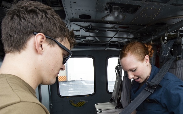 Two individuals are seated inside what appears to be a military helicopter or aircraft cabin. A young man wearing glasses and a tan t-shirt is assisting a woman in a navy blue flight suit with a seatbelt or harness. Through the window, a flight deck or tarmac surface is visible, suggesting the aircraft is aboard a naval vessel.
