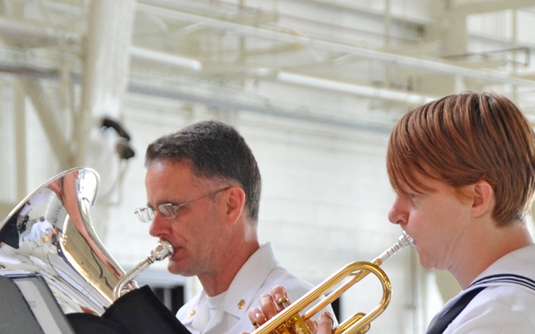 Two U.S. Navy musicians in uniform are playing brass instruments, reading from a music stand in what appears to be a hangar or large indoor facility. The person on the left is playing a silver flugelhorn or mellophone, while the person on the right, wearing a Petty Officer uniform with chevron insignia, is playing a gold/brass trumpet. Both are focused on their sheet music during what appears to be a formal military musical performance.