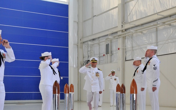 A formal U.S. Navy military ceremony is taking place inside a large hangar facility. A senior officer wearing a distinctive gold-braided white dress uniform and dark-brimmed cap walks along a red carpet while being rendered honors by sideboys standing at salute on both sides. The setting appears to be a modern aviation or naval facility with polished floors and large blue hangar doors.