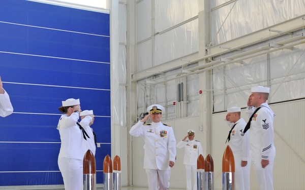 A formal U.S. Navy ceremony is taking place inside what appears to be a large hangar facility. A senior officer in dress whites walks along a red carpet while being saluted by a sideboy honor guard on both sides. A second officer in dress whites follows behind, also saluting.