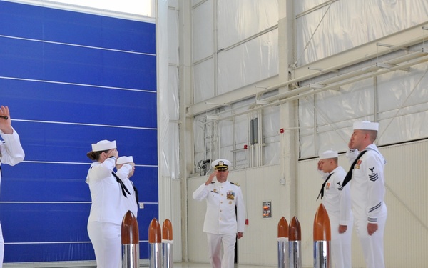 A formal U.S. Navy sideboy ceremony is taking place inside what appears to be a large hangar facility. A senior officer in dress whites walks along a red carpet while being saluted by a sideboy detail of enlisted sailors positioned on both sides. The setting features polished floors, blue hangar doors, and ceremonial bollard-style stanchions flanking the red carpet.
