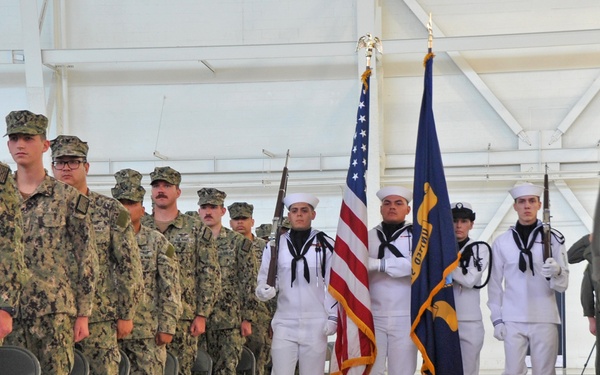 A military ceremony is taking place inside what appears to be a large hangar or indoor facility. A Navy color guard in dress whites is marching forward carrying the American flag and a Navy unit flag, while rows of personnel in camouflage uniforms stand at attention to the left. A person in a flight suit is visible on the right side of the frame.