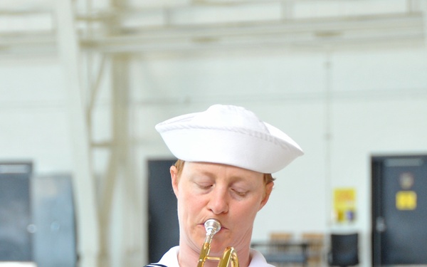 A U.S. Navy sailor in dress white uniform is playing a trumpet in what appears to be a large indoor facility such as a hangar or gymnasium. The sailor wears a traditional white Dixie cup hat and displays multiple service ribbons on their chest. The image is sharply focused on the subject with the background intentionally blurred.