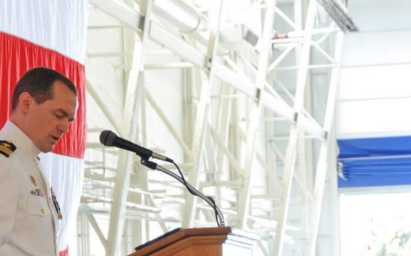 A naval officer in white dress uniform is speaking at a podium during what appears to be a formal military ceremony. The setting is inside a large hangar or similar facility decorated with red and white draping. A second uniformed naval officer stands at a secondary podium in the background.
