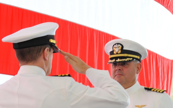 Two U.S. Navy officers in white dress uniforms are exchanging salutes during what appears to be a formal military ceremony, likely a change of command. The officer facing the camera displays multiple service medals, gold aviator wings, and captain-rank insignia on his shoulder boards. The backdrop consists of red and white draped fabric consistent with a formal naval ceremony setting.