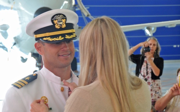 A military promotion or commissioning ceremony taking place inside an aircraft hangar. A woman is pinning insignia onto the uniform of a naval officer wearing a white dress uniform and cover with gold braid. Attendees in the background are photographing the moment.
