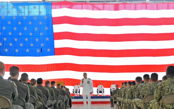 A formal military ceremony is taking place inside what appears to be a large aircraft hangar. A naval officer in white dress uniform stands at a podium or center aisle addressing rows of seated military personnel in various uniforms. A large American flag serves as the backdrop on the stage area.