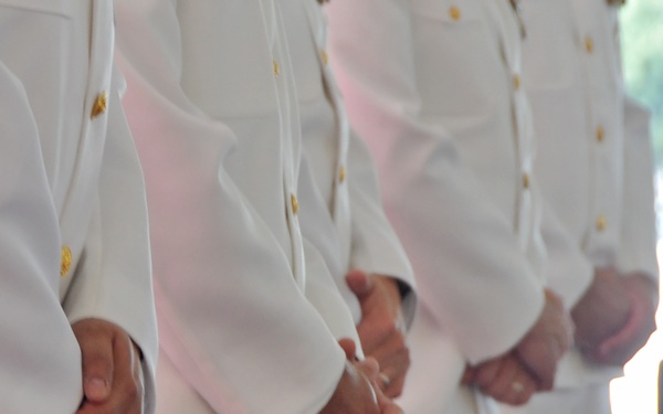 The image shows a row of individuals standing at attention in white military or naval dress uniforms. Their hands are clasped together in front of them at waist level, suggesting a formal ceremony or parade. The photo is taken from a torso-level perspective, focusing on the uniforms and hand positions.