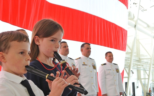 Two children are playing recorder flutes on a stage during what appears to be a formal military ceremony. Several naval officers in white dress uniforms stand in the background observing the performance. A large red and white draped backdrop is visible behind the group.