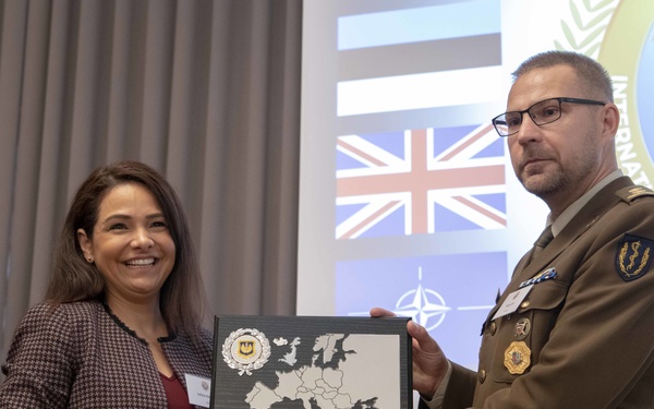 A woman in civilian attire and a military officer in olive/tan uniform are jointly holding a commemorative plaque featuring a map of Europe. The ceremony appears to be taking place at an international military conference or event, as indicated by the flags and organizational seal visible in the background. The setting suggests a formal award or recognition presentation.