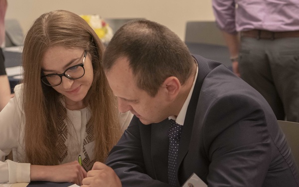 Two individuals are seated at a table covered with a blue tablecloth, collaborating over printed documents at what appears to be a professional conference or workshop. A young woman wearing glasses and a white patterned top is watching attentively as a man in a dark suit and tie writes or points to something on a document. Additional attendees and materials are visible in the background.