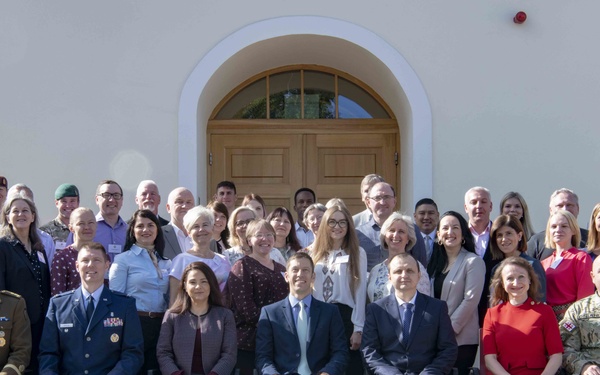 A large group of approximately 35-40 individuals posed for a formal group photograph in front of a white European-style building with an arched wooden door. The group includes both military personnel in various uniforms and civilians in business attire, suggesting an official multinational or interagency gathering. The front row is seated on chairs while subsequent rows stand on steps behind them.