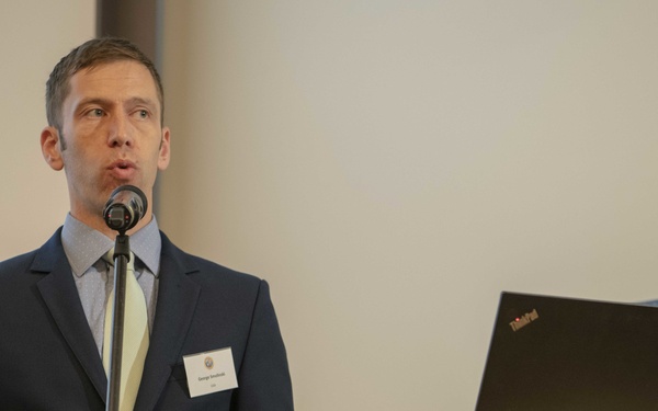 A man in a dark navy suit with a light tie is delivering a presentation at a formal conference or seminar. He stands at a microphone stand and has a name badge on his lapel. A ThinkPad laptop sits on a round podium table to his right, and a projection screen with a black-and-white image is visible behind him.