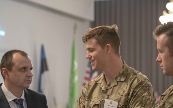 Three men are engaged in conversation at what appears to be a formal military or diplomatic event. Two of the men are wearing British Army MultiTerrain Pattern (MTP) camouflage uniforms with Union Jack patches, while the third is dressed in civilian business attire with a name badge. The setting appears to be a conference or reception hall with multiple national flags visible in the background.