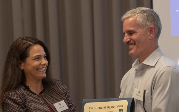 A woman is presenting a Certificate of Appreciation to a man named Major Garlisi during what appears to be a formal military or professional symposium. Both individuals are smiling and shaking hands while jointly holding the framed certificate. The event appears to be the first International Military Rehabilitation Symposium held in Tallinn, Estonia, in June 2019.