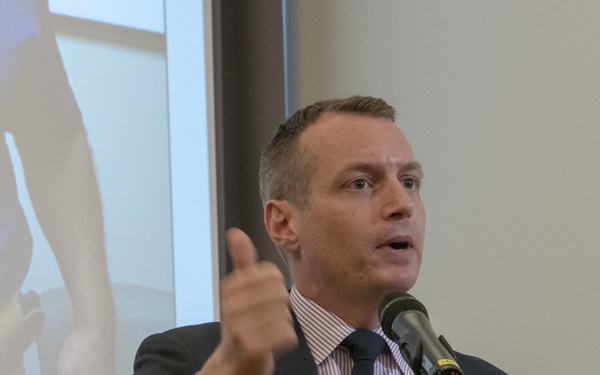 A man in a dark suit is speaking at a podium microphone, gesturing with his right hand in a thumbs-up or pointing motion. He is wearing a name badge on his lapel and a striped dress shirt with a dark tie. A presentation slide is visible on a projection screen behind him.