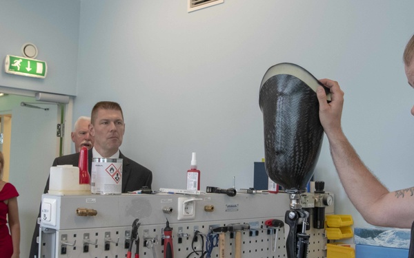 A bearded man in a black t-shirt is presenting or demonstrating a carbon fiber prosthetic socket to a small group of observers in what appears to be a prosthetics workshop or rehabilitation facility. The workbench behind him displays various prosthetic limb components, tools, and assembly equipment on a pegboard. The setting suggests a professional demonstration or tour of a prosthetics manufacturing or fitting facility.