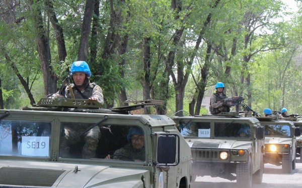 A convoy of military HMMWV (Humvee) vehicles travels along a tree-lined road. Soldiers wearing distinctive light blue helmets, indicative of United Nations peacekeeping forces, are visible manning the vehicles, some standing through roof hatches with weapons. The lead vehicle bears the license plate '7287 KA' and a placard marked 'SE19'.
