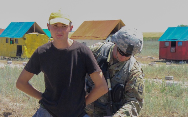 A soldier in full combat gear and camouflage uniform is conducting a pat-down or search of a civilian male wearing a dark t-shirt, yellow cap, and gray sweatpants. The setting appears to be a military training area with colorful small structures or mock buildings in the background. This appears to be a training exercise based on the controlled environment and mock village setup.