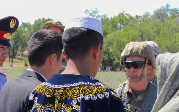 The image shows a group of people engaged in what appears to be a civil-military interaction or community engagement meeting outdoors. The group includes Western military personnel in combat uniforms and helmets, a local police or military officer in a blue uniform with a red-banded cap, and civilians wearing Central Asian traditional attire including an embroidered chapan robe and a white skullcap (doppa/tubeteika). The setting appears to be a rural or semi-rural area in Central Asia, likely Kyrgyzstan, Kazakhstan, Uzbekistan, or a neighboring country.