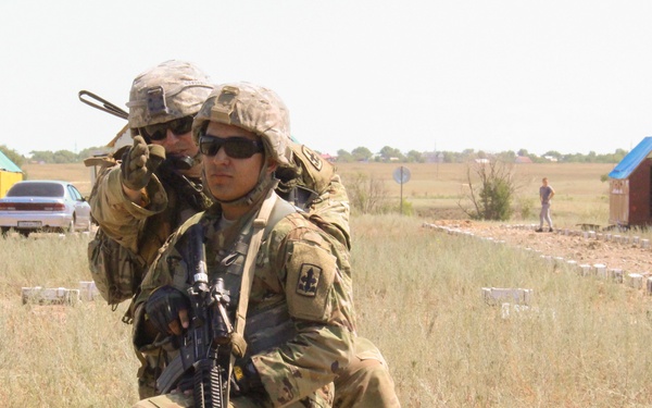 Two soldiers in U.S. Army MultiCam/OCP camouflage uniforms and ACH helmets are conducting what appears to be a military training exercise in an open, arid field. One soldier is kneeling and holding an M4 carbine rifle while the other stands behind him pointing in a direction, suggesting a tactical instruction or coordination moment. The background features colorful small structures (likely target houses or training props) arranged across a flat, semi-arid landscape.