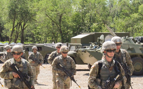 A group of U.S. Army soldiers in full combat gear are marching across a gravel area during what appears to be a military training exercise or field operation. The soldiers are equipped with assault rifles, body armor, helmets, and tactical gear. Several military vehicles, including armored personnel carriers and HMMWVs, are visible in the background.