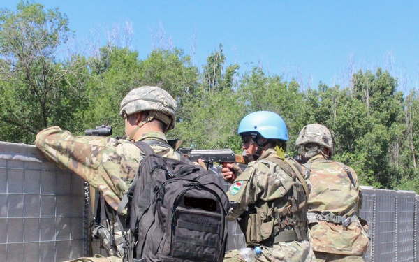 Three soldiers are positioned behind a HESCO barrier wall in what appears to be a military training exercise. One soldier wears a distinctive UN-style blue helmet, while the others wear standard camouflage combat helmets. They are in tactical crouching positions with weapons raised, suggesting a simulated urban or perimeter defense drill.