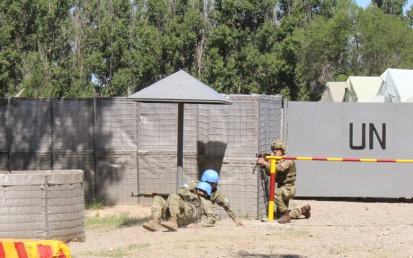 The image shows what appears to be a United Nations peacekeeping training exercise at a simulated base or checkpoint. Two soldiers wearing distinctive UN blue helmets are in tactical positions near a gate barrier, with one prone and one kneeling while holding rifles. The scene includes UN-marked structures, red-and-yellow painted concrete barriers, and military tents in the background.