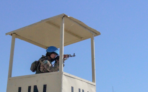 A United Nations peacekeeper is positioned in a white guard/observation tower marked with 'UN' on two visible sides. The soldier is wearing a distinctive UN blue helmet and is aiming a rifle outward from the elevated post. The tower appears to be reinforced with sandbags or HESCO barriers at its base.