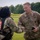 Two U.S. Army soldiers in OCP (Operational Camouflage Pattern) uniforms are shaking hands on a grassy field. The soldier on the right, identified by name tape as 'MAXWELL,' is wearing a U.S. Army uniform and holding a smartphone. The soldier on the left is wearing a parachute harness with visible orange emergency handle, suggesting a airborne/parachute activity has recently taken place.