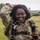 A female U.S. Army soldier in Operational Camouflage Pattern (OCP) uniform is smiling broadly while standing on a grassy field. She is wearing a parachute harness, suggesting she has recently completed or is preparing for a airborne/paratrooper activity. She appears to be in a relaxed, celebratory mood, holding small objects in one hand.