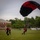 A parachutist is landing on a green athletic field with a black and red parachute canopy still inflated overhead. Two ground crew members dressed in matching black and red uniforms are approaching to assist with the landing. The setting appears to be a military or academic campus based on the brick building and bleachers visible in the background.