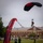 A tandem parachute jump is captured moments before landing on a grassy field at what appears to be a military installation. A black and red parachute canopy is visible overhead as two jumpers descend together, with a brick building labeled 'Hays Hall' bearing a 10th Mountain Division patch visible in the background. A promotional banner reading 'Black Daggers' — the U.S. Army's parachute demonstration team — is prominently displayed in the foreground.