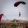 A tandem parachute jump is taking place over a military installation, with two jumpers descending under a black and red parachute canopy. The landing zone is a grassy field in front of a brick building identified as 'Hays Hall,' which displays the 10th Mountain Division insignia. Spectators and ground crew observe the landing from the field below.