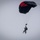 Two individuals are performing a tandem skydive, descending under a black and red rectangular parachute canopy against an overcast gray sky. The parachute appears fully deployed and inflated, suggesting a controlled descent. One person is strapped to the front of the other, characteristic of a tandem jump configuration.