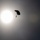 A silhouetted parachutist is captured mid-descent against a bright, hazy sun in an overcast sky. The strong backlight creates a dramatic contrast, rendering both the parachute canopy and the suspended figure as dark silhouettes. The image has a moody, atmospheric quality due to the diffused sunlight and gray sky.