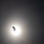 A silhouetted paraglider or parachutist is captured against a bright, overexposed sun in an otherwise dark, hazy sky. The subject appears as a dark shape with a canopy visible above a suspended figure. The strong backlight creates a dramatic contrast between the glowing solar disc and the dark silhouette.