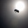 A silhouetted parachutist is captured mid-descent against a bright, hazy sky with the sun serving as a strong backlight. The image is shot in a high-contrast, backlit style that renders the parachute canopy and jumper as dark silhouettes. The overcast or hazy atmospheric conditions diffuse the sunlight, creating a glowing orb effect in the upper right portion of the frame.