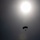 A silhouetted parachutist descends against a bright, overcast sky with the sun visible as a glowing orb near the top center of the frame. The image is captured in a backlit, high-contrast style, rendering the parachute and jumper as dark silhouettes. Lens flare artifacts are visible in the lower portion of the image.