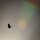 A silhouetted parachutist descends against an overcast sky featuring a visible rainbow arc in the upper right portion of the frame. The parachute canopy is fully deployed and appears as a dark crescent shape against the muted, grey-toned sky. A secondary optical phenomenon, possibly a sun dog or iridescent cloud patch, is visible as a warm orange-yellow glow near the center-right of the image.