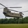 A United States Army CH-47 Chinook tandem-rotor helicopter is hovering at low altitude over a green grassy field. The aircraft is painted in olive drab and displays 'UNITED STATES ARMY' markings on its fuselage. The setting appears to be a rural or training area surrounded by dense green tree lines under an overcast sky.