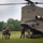 A military CH-47 Chinook helicopter with tail number 14-08451 is landed on a grassy field. Four individuals, some wearing red jackets and tactical gear with backpacks, are gathered near the rear ramp of the aircraft. The scene appears to depict a personnel loading or briefing operation in a rural outdoor setting.