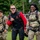 Three individuals wearing parachute harnesses are walking on a grassy field, appearing to be after a skydiving or airborne jump activity. Two are wearing U.S. Army camouflage uniforms with what appears to be 10th Mountain Division patches, while one person in the center wears a red and black civilian jumpsuit. The group appears in high spirits, with two of them smiling broadly and one giving a 'shaka' hand gesture.