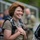 A woman with short red hair is smiling broadly while wearing a parachute harness and holding clear protective goggles against her chest. She appears to be at a skydiving or airborne activity event, surrounded by other participants in the blurred background. Her expression conveys excitement or satisfaction, likely post-jump or pre-jump.