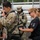 A military parachute equipment check is underway, with a civilian or instructor in a black 'Airborne' t-shirt inspecting the harness of a soldier wearing 10th Mountain Division patches and a parachute rig. Multiple soldiers in OCP (Operational Camouflage Pattern) uniforms are visible in the background near bleachers, suggesting a pre-jump staging area. The scene appears to be an airborne operations preparation event.