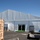 The image shows a large white temporary structure or event tent set up in what appears to be a paved parking lot or industrial area. The tent is a professional-grade clearspan structure with rigid framing and white PVC sidewalls, featuring a glass double-door entrance with a green artificial turf mat. The surrounding area includes shipping containers, utility equipment, light poles, and various cables and hoses running along the base of the structure.