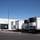 The image shows a parking lot setup with multiple white refrigerated trailers parked alongside a large white temporary structure or tent. The trailers appear to be connected to power or utility lines running along the ground. A worker in a high-visibility vest is visible near the tent structure on the left side.